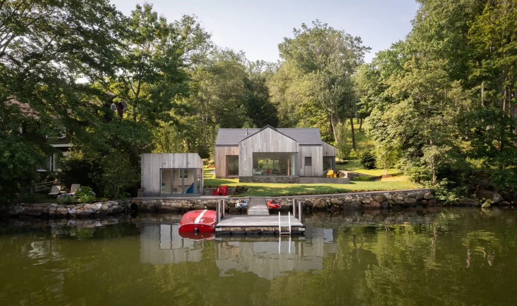 Single story wood clad passive house sits on grassy lawn overlooking lake.