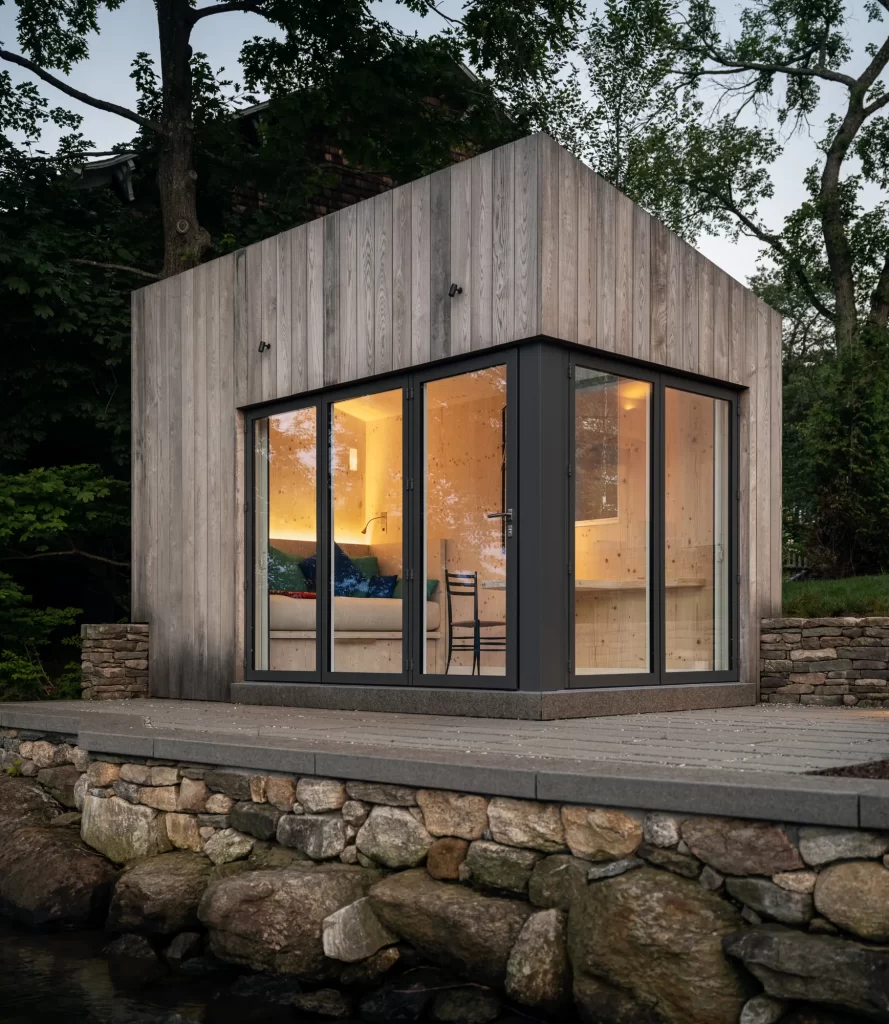 Small wood clad building with large glazed doors sitting on deck overtop of stone wall.