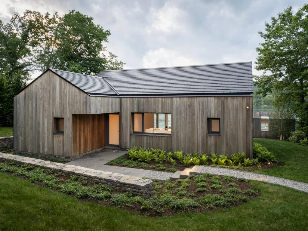 View of entrance to single story wood clad house with Tesla solar roof, surrounded by ferns, shrubs and green grass lawn.