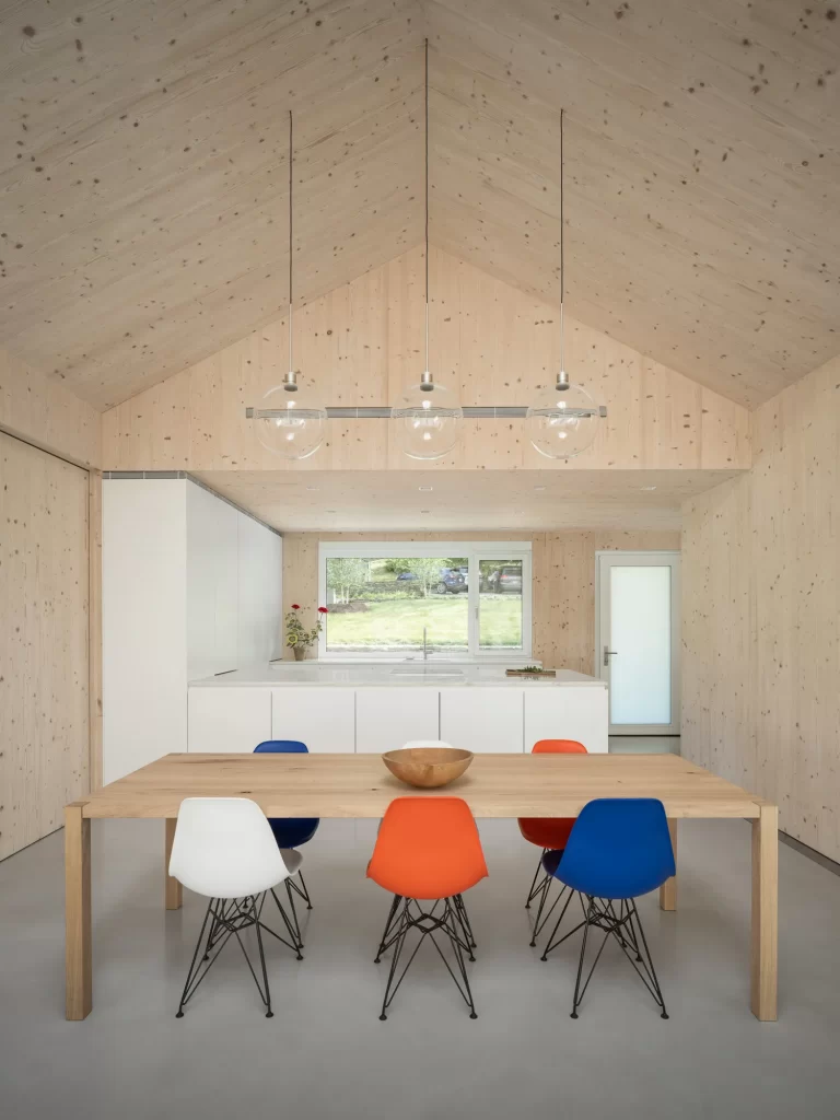 Red, white and blue chairs sit at simple wood table with bright white kitchen behind, interior walls and ceiling are warm cross-laminated timber (CLT) wood.