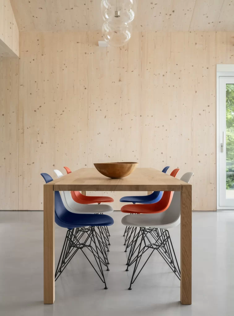Red, white and blue chairs sit at a simple wood table surrounded by cross-laminated timber (CLT) walls and concrete floors.