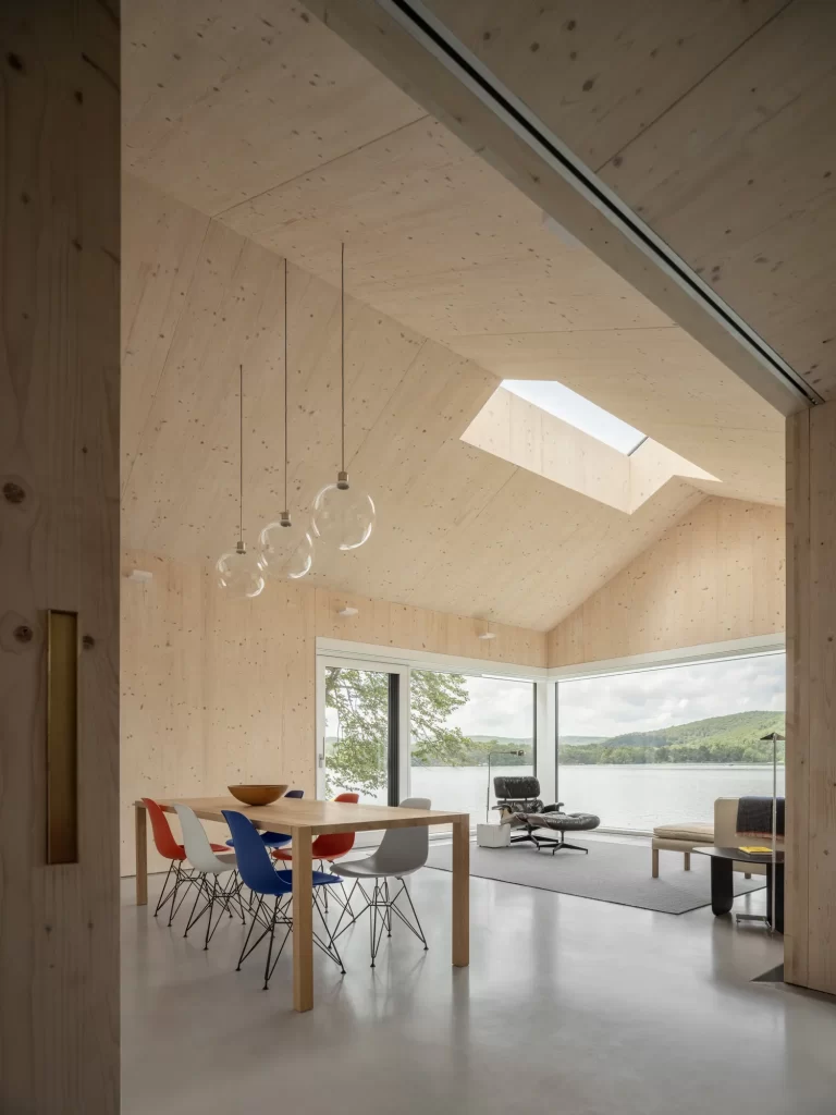 Warm cross laminated timber wood interior walls and ceilings, cement flooring and large glazing surround modern furniture overlooking a lake view.