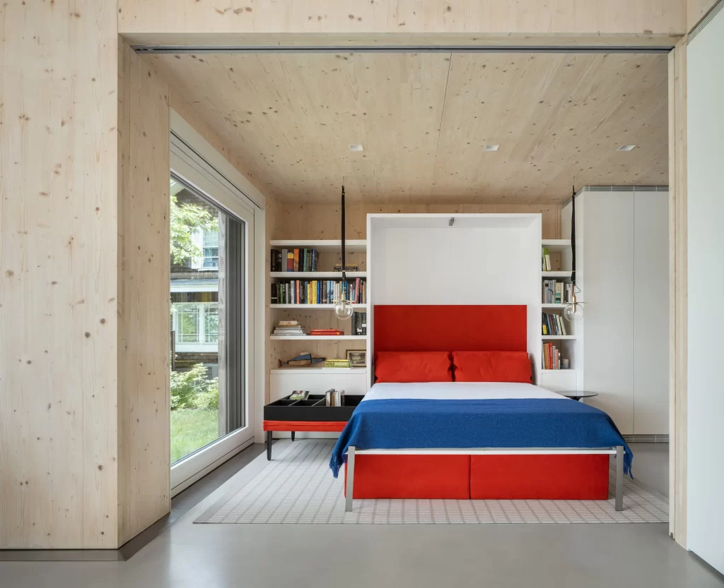 Cross Laminated Timber Interior walls and ceilings and large triple paned glass door frame a bright red and blue Murphy bed in this modern living space..