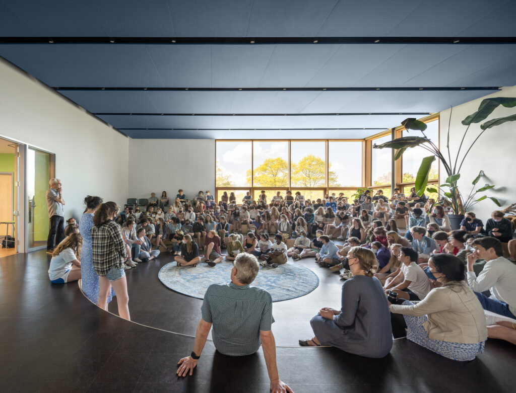 A large light filled room with huge curtain wall windows on two sides is filled with students sitting on the steps for the "All School Meeting".